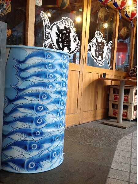 居酒屋の前にさかなの大軍が描かれたドラムカン